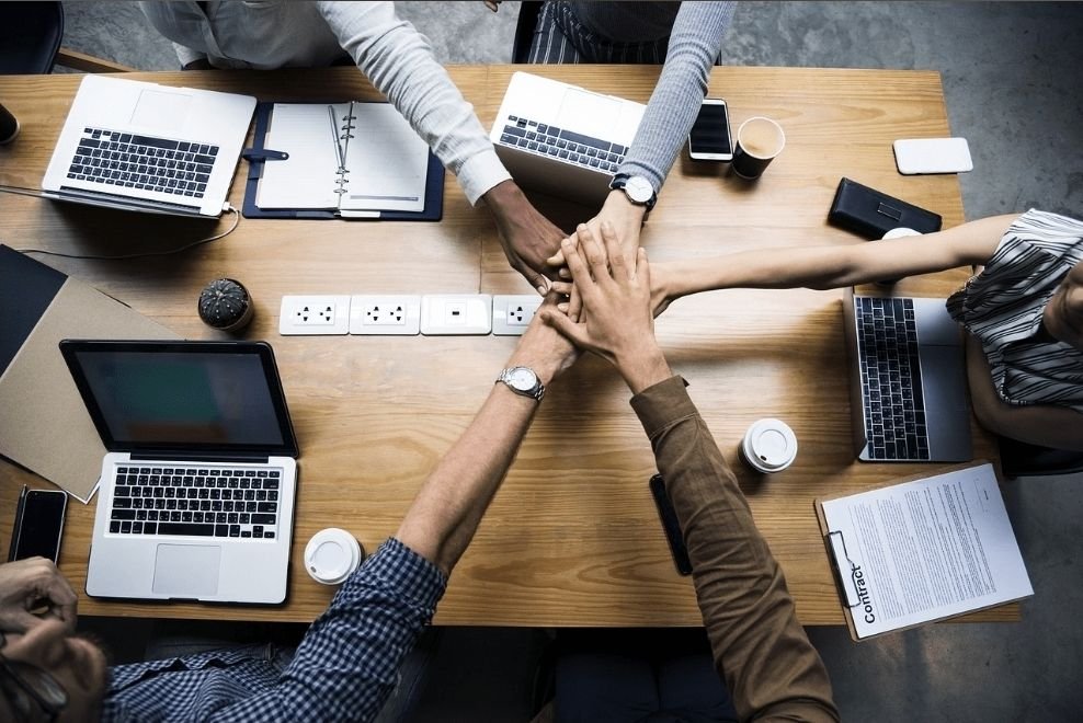Team joining hands over a table of laptops and plans